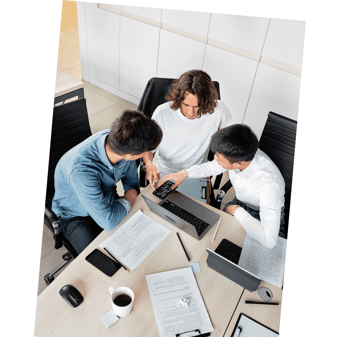 Three people sitting around a table with laptops and documents in a collaborative office environment.