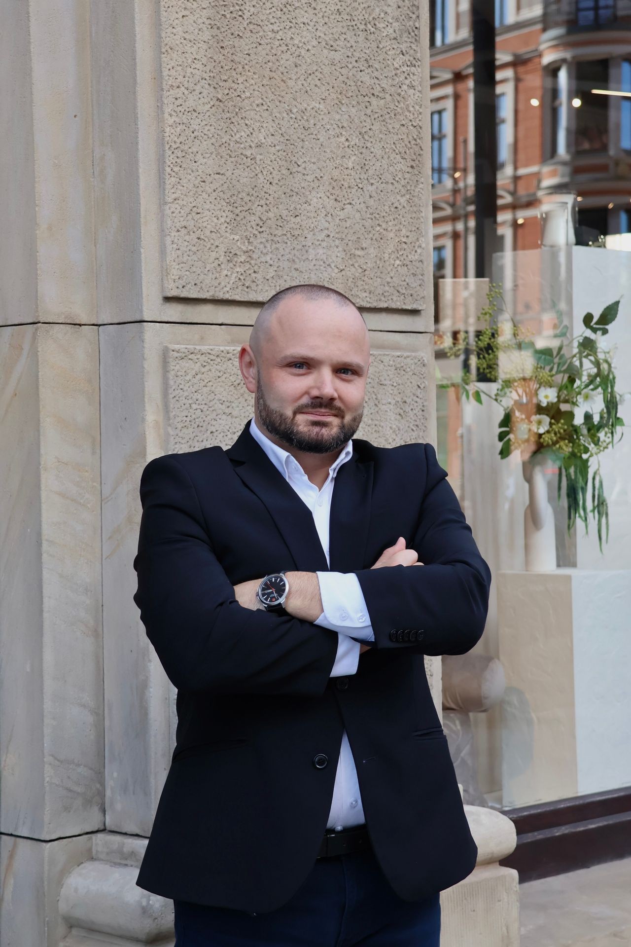 Man in black blazer stands confidently with arms crossed, building and plants in background.