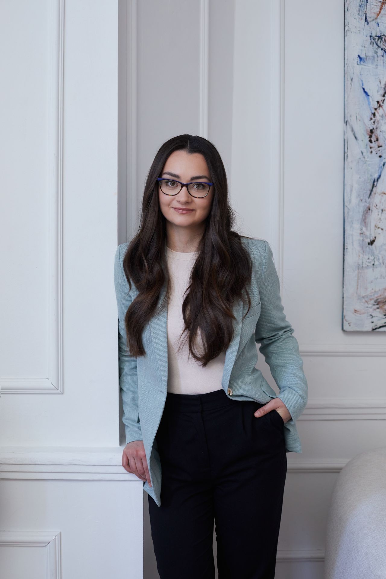 Woman with long hair and glasses wearing a blazer, leaning against a wall in a modern room.