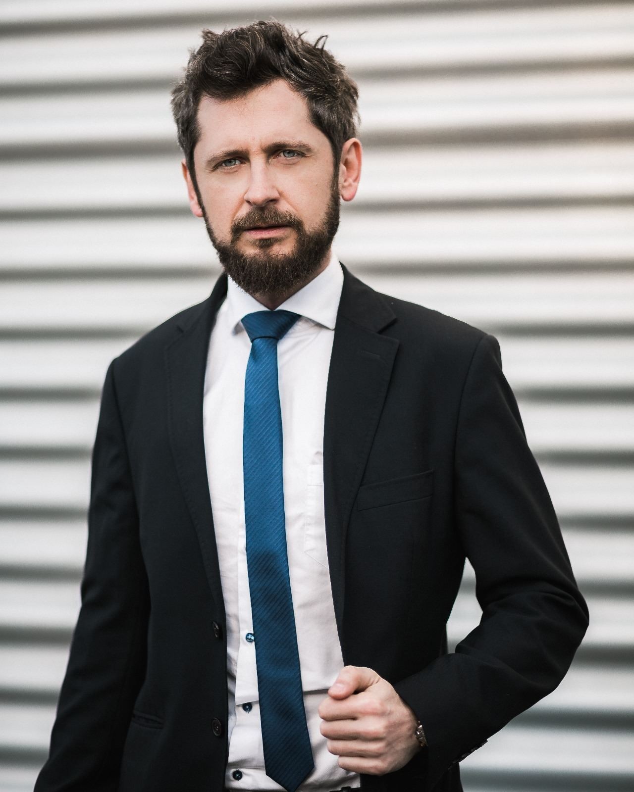 Man in a black suit and blue tie standing in front of a metal shutter background.